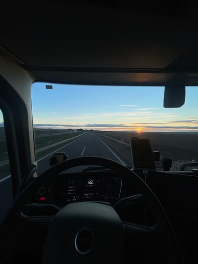 The image depicts a vehicle's interior view while driving on a highway during sunrise. The sun is visible on the horizon, and the sky transitions from blue to orange near the horizon. The dashboard, steering wheel, and a display showing "82" are in the foreground. The highway stretches straight ahead, and contrails from a plane are noticeable in the sky.