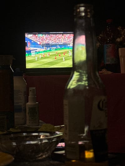 A bottle of alcohol is placed in front of a television screen displaying a soccer match. The screen shows a game in progress, with players in red and white uniforms on a green field. The score is visible on the screen, indicating 1-1 in the 31st minute. The setting appears to be indoors, possibly a living room, with a dark background. Other objects visible include a red cloth and some unidentifiable items on a surface behind the television. A hand-sanitizer bottle is also on a surface. The lighting suggests it might be evening or nighttime. The event is a soccer match being watched on television, possibly during a gathering or while relaxing at home. The overall mood is casual and relaxed.