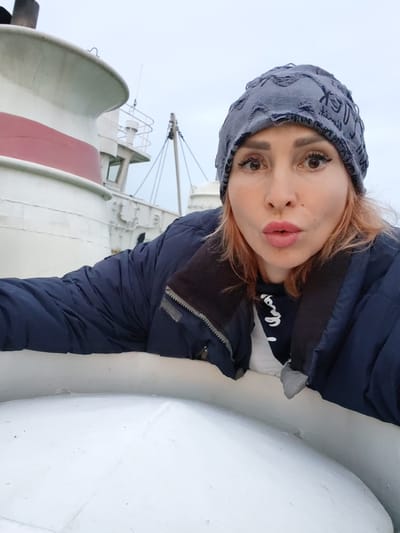 A woman in a gray beanie and navy jacket is taking a selfie on the deck of a ship. The ship has a white hull with a red stripe. The sky is overcast. The woman is looking at the camera with her lips puckered. She has light brown hair and dark brown eyes. The image was taken in Ivanov Bor, Russia.