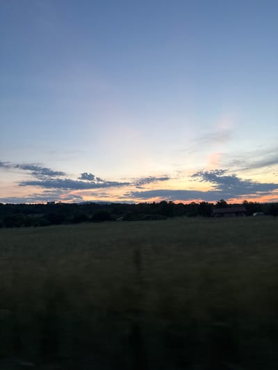 The image depicts a sunset over a rural landscape near the city of Slokoshtitsa, Bulgaria. The sky is a gradient of blue transitioning to orange and pink near the horizon, with scattered clouds. Below the sky, a dark, tree-lined horizon stretches across the middle ground. In the distance, silhouetted structures, possibly buildings, are visible amidst the trees. A small, dark structure, resembling a shed or small house, is present on the right side of the image, partially obscured by trees. The foreground consists of a dark, blurry field, suggesting it is either very late in the day or the image was taken from a moving vehicle. The overall scene is tranquil and evokes a sense of the countryside at dusk.