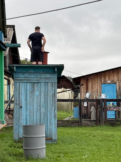 A man stands on top of a blue wooden outhouse in what appears to be a rural setting in the city of Gonja, Russia. The man, seen from the back, is wearing a dark t-shirt and dark shorts. He has his hands on his hips. The outhouse is weathered and has a hinged door on the front. A grey metal barrel sits on the grass in front of the outhouse. In the background, there is a wooden building with a blue door and a fenced yard containing several metal buckets. The sky is overcast.