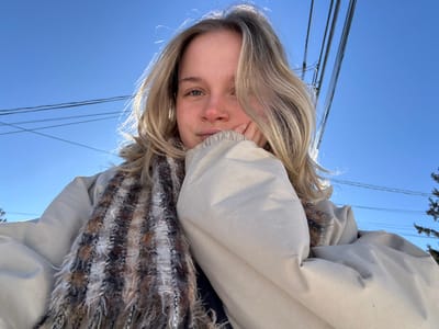 A close-up outdoor selfie of a female subject, taken in Kingston, Canada, under a clear blue sky. The woman, with shoulder-length blonde hair and a small ring piercing in her left nostril, looks directly at the camera with a neutral expression. She wears a light beige padded jacket and a textured scarf, displaying shades of brown, black, white, and gray. Her left hand, with fingers curled, supports her chin. The background consists of a bright blue sky, transversed by multiple dark utility wires. A dark green conifer tree is partially visible in the lower right, and strong sunlight from behind the subject creates a halo effect on her hair.