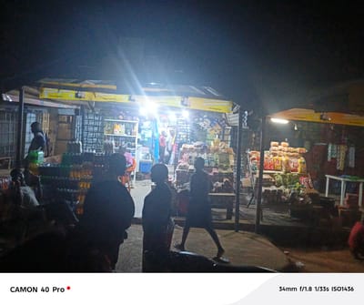Captured at night in Langtang, Nigeria, the image presents an outdoor market scene. The foreground is dominated by silhouetted figures of individuals standing and walking. The market itself features stalls laden with a variety of goods, including stacked bread, bags of food items, and beverages. Artificial lighting illuminates the market area, creating a contrast with the darker surroundings. Banners with the slogan "CHOOSE HAPPINESS" are visible above the stalls. In the background, people are seated and interacting, while others are browsing the market's offerings. The general atmosphere suggests a lively commercial activity at night.