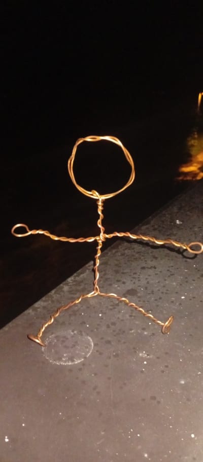The image shows a wire figurine resembling a stick figure, made of copper-colored wire, positioned on a dark, speckled surface, possibly a ledge or countertop. The figurine has a circular head, a body formed by twisted wire, outstretched arms with looped ends, and legs bent at the knees with looped feet. The background is dark, with faint reflections of light, suggesting it is nighttime. The figurine is the central focus of the image. There is no indication of the city of Karviná, Czechia, in the image.