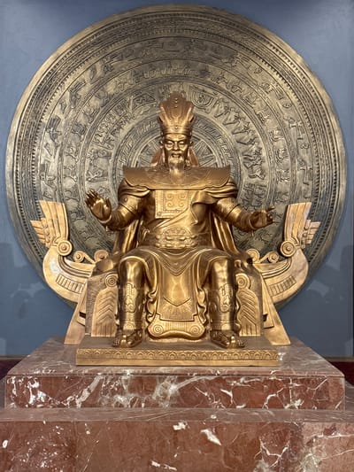 A full shot shows a golden statue of a king or emperor seated on a throne. The statue is positioned in front of a large, circular bronze disc decorated with intricate patterns and figures. The throne itself has a boat-like structure on either side. The statue is wearing a crown, ornate armor, and flowing robes. The king has a beard and is gesturing with his hands. The statue stands on a multi-tiered marble pedestal. The background is a plain blue wall. This statue is located at the Hung Kings' Temple complex in Hy Cương, Vietnam.