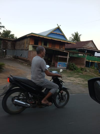 A man is riding a black motorcycle on a road in Pallawarukka, Indonesia. The man is wearing a grey t-shirt and grey pants. The motorcycle has a black seat and a silver exhaust pipe. In the background, there are several buildings with wooden walls and colorful roofs. One of the buildings has a sign that says "ES TELUR ES". There are also palm trees in the background. The sky is clear and the sun is setting.