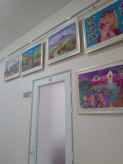 The image shows a hallway in Nor Hachin, Armenia, with several framed paintings displayed on a light-colored wall. To the left, there is a sign with Armenian text. A white door with a frosted glass panel and a silver handle is visible in the center-right of the image. Above the door, a small white sign with the number "13" is affixed. The paintings appear to be folk art or child-like drawings, depicting various scenes including landscapes with mountains and flowers, a woman with a traditional headdress, and a surreal scene with an eye and figures. The overall impression is of an indoor public space, possibly a school, clinic, or community center.
