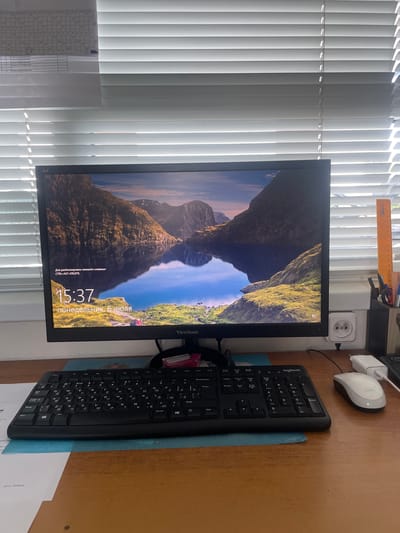 The image shows a computer setup on a wooden desk. A large monitor displays a scenic landscape of mountains and a lake, with the time "15:37" and the date "понедельник, 6 июля" (Monday, July 6) visible. Below the monitor is a black keyboard with both Cyrillic and Latin characters. A white computer mouse is to the right of the keyboard, and a white power adapter is plugged into an electrical outlet on the wall behind the monitor. The scene is illuminated by sunlight coming through horizontal blinds on a window. To the left of the monitor, a printed schedule or calendar is partially visible, attached to the blinds. To the right of the monitor, a white pencil holder contains pens, scissors, and an orange ruler. The image does not contain enough information to determine the location in the city of Salsk, Russia.