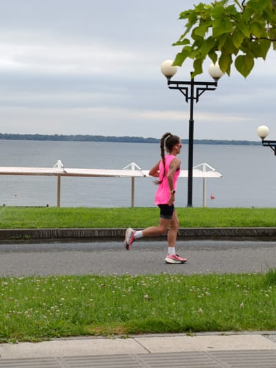 A young woman with a braid runs along a paved path next to a body of water. She is wearing a bright pink tank top and black shorts. The path is bordered by a strip of green grass with small white flowers and a row of streetlights. In the background, a calm body of water stretches to the horizon, with a distant treeline visible. The sky is overcast. The scene appears to be a recreational area within Voskresensky District, Russia, likely a waterfront park or promenade.
