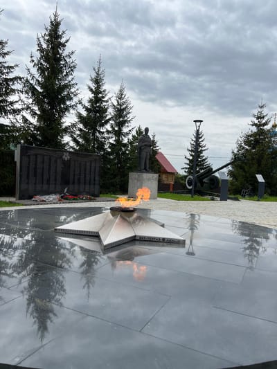 The image shows a war memorial in Введенская Слобода, Russia. The memorial features an eternal flame, a statue of a soldier, and a black granite wall inscribed with names. A cannon is also present. The ground around the memorial is dark and reflective, with trees in the background and a cloudy sky overhead.