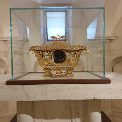 The image displays a human skull contained within an ornate reliquary, secured within a clear glass display case. This setup is located in the Crypt of San Ciriaco inside the Cattedrale di San Ciriaco in Ancona, Italy.

The reliquary is predominantly gold-colored with silver-toned accents on its base and decorative elements. It features intricate Baroque-style ornamentation, including scrolling foliage, putti (cherubic figures), and floral motifs. A red fabric element is visible on the reliquary's lid, positioned beneath a golden, cross-shaped or X-shaped finial. The human skull is clearly visible through an oval opening on the reliquary's front. The glass display case, which encloses the reliquary, rests on a dark brown rectangular base. This entire arrangement is placed atop a large, light-colored stone pedestal or altar.

The immediate environment is an interior space characterized by light-colored stone architectural features, including prominent columns with classical capitals and arched structural elements in the background. A high, rectangular window provides ambient, indirect illumination. No individuals are present, and no actions or interactions are depicted within the scene.