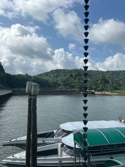 The image depicts a waterfront scene in Pak Nam, Thailand. In the foreground, a decorative metal chain with cup-like segments hangs vertically, obscuring part of the view. Two thick, rough-barked wooden posts, topped with two weathered, cylindrical structures, stand in the water to the left.
Moored in the water are several white boats with dark trim and tinted windows. One boat in the foreground has a white canvas cover, while another boat behind it features a green arched canopy. The water appears calm and slightly rippled.
In the background, a body of water extends towards a lush, green shoreline covered in dense tropical vegetation and palm trees. A low-lying building with a covered walkway or pier is visible on the left side, extending over the water. Further inland, a gently sloping hill is densely forested. The sky above is a mix of blue sky with scattered white cumulus clouds. The overall impression is a serene tropical setting with nautical elements.