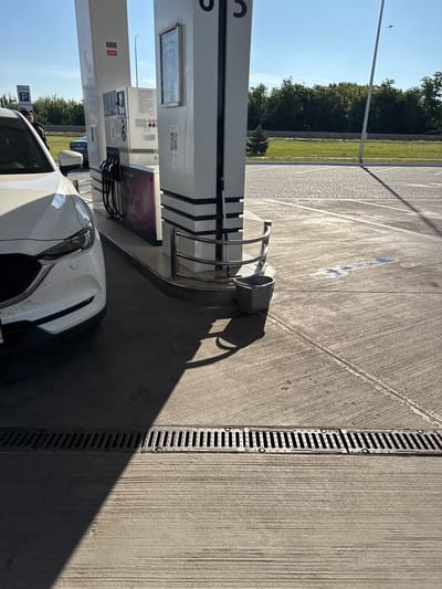 A white car is parked at a gas station in Nerubaiske, Ukraine. The car is partially visible on the left side of the image, with its headlights and front bumper in focus. The gas pump is tall and white, with multiple hoses and screens. A dark grey bucket is placed on the ground near the pump. The ground is concrete, with a drainage grate running horizontally across the bottom of the image. The sky is blue and clear, with some trees in the background. The sun is shining, casting shadows on the ground.