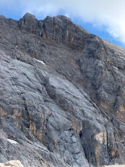 The image shows a steep, rocky mountain face under a bright, partly cloudy sky. The rock is grey and textured, with visible layers and crevices. In the distance, a thin cable stretches across the sky, likely part of a cable car system. Several small figures, appearing to be people, are visible on a narrow ledge or path carved into the mountainside. They are too small to discern details about their clothing or any specific activities.
The lighting suggests it is daytime, with the sun casting shadows that highlight the rugged terrain. The presence of some snow patches on the rock indicates a cool or high-altitude environment. The overall scene evokes a sense of scale and adventure, with the vastness of the mountain contrasted against the small human figures. There is no visible text in the image. The scene is likely a hiking or climbing area, with the cable car suggesting a tourist or recreational destination. The location context of Wildermieming, Austria, places this scene within the Alps, known for dramatic mountain landscapes and outdoor activities.