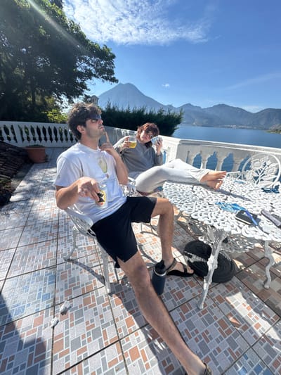 Two individuals are seated on a tiled patio overlooking Lake Atitlán and surrounding mountains, including a prominent volcano, under a clear blue sky in San Pablo La Laguna, Guatemala. The patio floor features geometric brown, grey, and blue tiles, bordered by a white ornate balustrade.

A male individual, with dark hair and wearing sunglasses, a white t-shirt with an avocado graphic, black shorts, and dark sandals, is seated in a white ornate metal chair on the left. He holds a "Gallo" brand beer can in his right hand and has his left index finger raised near his temple. A dark water bottle is positioned on the floor near his feet.

A female individual, with reddish-brown hair and wearing a grey long-sleeved top, grey sweatpants, and sunglasses pushed onto her head, is seated in a matching white ornate metal chair on the right. Her bare feet are propped on a white ornate metal table. She holds a clear glass with an amber liquid in her left hand and a fanned hand of playing cards in her right hand. On the table, several playing cards are scattered, along with a small dark notebook and a pen.

Behind the balustrade, green trees and bushes are visible. The lake extends into the mid-ground, with multiple mountains and a distinct conical volcano dominating the distant horizon. Bright sunlight casts strong shadows across the patio. Two small crumpled white objects are on the tiled floor near the male's left foot.