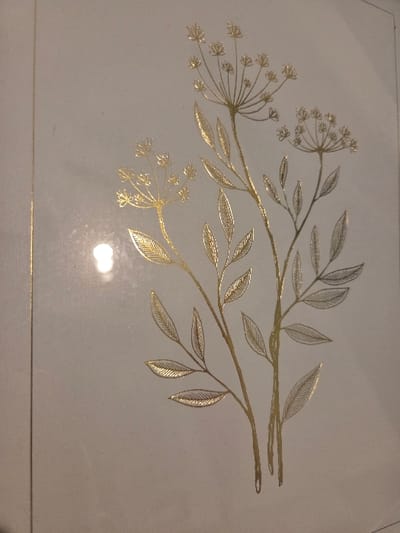 The image displays a gold-foil botanical illustration on a light-beige surface. Three stylized plant stems rise from the bottom, diverging upwards. The tallest central stem features multiple elongated leaves and a prominent, radially structured flower cluster. To its left, a shorter stem bears leaves and a smaller flower cluster. A third stem, to the right, displays leaves and another flower cluster. All illustrated elements are metallic gold, exhibiting a reflective sheen. A bright, localized glare from a light source is visible on the left side, slightly below the image center. The background is plain. No individuals or active subjects are present. The scene is located in Blainville-sur-Orne, France.