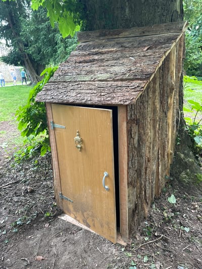 This image shows a rustic, handcrafted wooden shed, approximately human-sized, leaning against the base of a large tree. The shed is constructed from rough-hewn logs and bark, with a shingled roof made of bark. A single wooden door, slightly ajar, reveals a dark interior. The door has a silver handle and a brass knocker shaped like a lion's head. The shed appears to be situated outdoors in a grassy area with scattered leaves and branches on the ground, suggesting a natural or park-like setting. In the background, several individuals are walking on a path, and further in the distance, a stone structure is visible. The overall impression is of a whimsical or fairytale-like structure integrated into its natural surroundings.