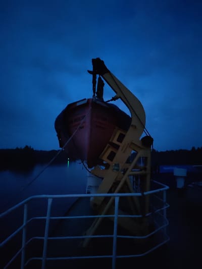 An orange lifeboat is suspended by a yellow davit on the deck of a ship at dusk. The sky is a deep blue, with some faint clouds. The water is dark, reflecting the dim light. A white railing is visible in the foreground, leading away from the lifeboat and davit. Trees are silhouetted against the horizon in the distance. The scene appears to be located in or near Badozhskoi Pogost, Russia, as indicated by the text on the lifeboat which reads "БДОЖСКОЙ ПОГОСТ". There are two figures standing near the lifeboat, but their features are not clearly discernible in the low light. The lifeboat is connected to the davit by ropes. A faint green light is visible in the water below the lifeboat.