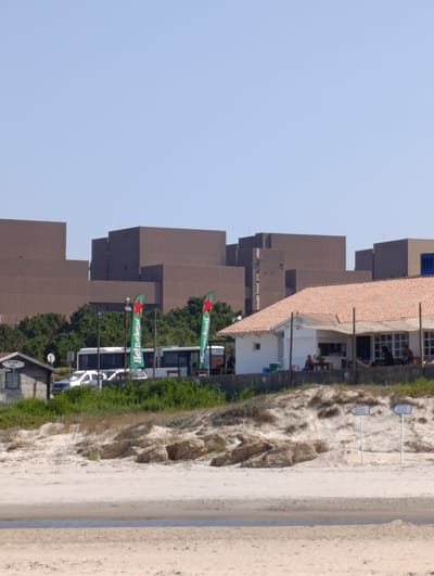 The image shows a beach scene in Sines, Portugal, with a cafe or restaurant in the foreground. The cafe has a tiled roof and an outdoor seating area where several people are visible. Behind the cafe, there are two flags with the Heineken logo. Further in the background, there is a white bus and some cars parked. Beyond the bus and cars, a line of lush green trees separates the beach area from a series of large, brown, modern buildings with flat roofs. The sky is clear and blue. The sand in the foreground is light-colored and appears to be a typical beach environment. There are also some large rocks scattered on the sand. Two directional signs are visible on poles in the middle ground.