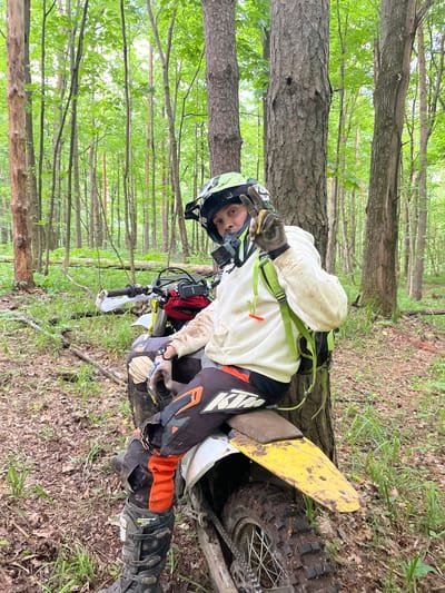 A motorcyclist wearing protective gear sits on a dirt bike in a wooded area. The rider wears a helmet, gloves, and boots. The dirt bike appears to be positioned next to a tree. The surrounding environment consists of trees and ground foliage. The location is undetermined and cannot be identified as Gruzinskii, Russia.