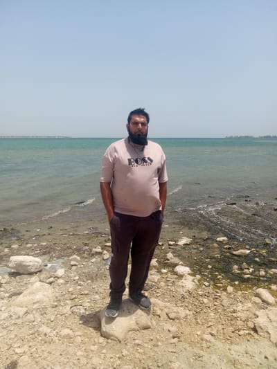 A man with a beard stands on a rocky beach on the coast of the Persian Gulf in Al `Aziziyah, Saudi Arabia. He is wearing a light pink t-shirt with "BOSS NEW STYLE" printed on it, along with dark brown pants and grey sneakers. The sea is a light turquoise color with visible waves. The sky is pale blue. The foreground consists of rocks and algae. In the distance, a low-lying landmass is visible across the water. The man is standing with his hands in his pockets.