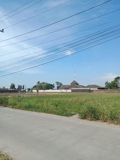The image shows a landscape in Cangkring, Indonesia, under a clear, blue sky. Overhead are a network of power lines. In the foreground is a gray concrete road with grassy verges. Beyond the road, there's a field of tall, green grass and weeds. In the distance, a low wall surrounds a building complex featuring a multi-tiered roof structure, topped by a radio or telecommunications tower. Additional buildings are visible adjacent to the main structure. Trees are interspersed around the buildings. In the far background, there is a hint of other structures and trees.
