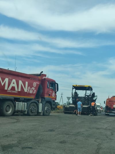 In Beya, Russia, a construction scene unfolds under a partly cloudy sky. A large red MAN dump truck is parked on an asphalt road. On the side of the dump truck, the word "MAN" is prominently displayed in large white lettering, along with the text "[Greshnik]" below it. Further details on the truck include a logo with Cyrillic text and a sticker of cartoon characters.

To the right of the dump truck, a VÖGELE asphalt paver is positioned, with several individuals working around it. One person, wearing an orange vest, stands near the paver. Another person in a blue shirt and shorts is also present, along with two others in darker clothing. A smaller yellow construction vehicle is partially visible behind them.

In the background, other vehicles are present, including a tanker truck with Cyrillic text and a license plate that reads "M 298 B X 50". Power lines stretch across the horizon. The ground is covered in asphalt and some debris, suggesting ongoing road construction or repair.