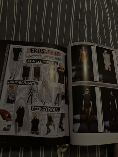 An open magazine rests on a striped surface. The pages showcase fashion photography and illustrations. The right page features a series of illustrated fashion designs with labels in Italian such as "Nerosunudo," "Corpobarrato," and "Clergydoll." The adjacent page has a grid of black and white photographs of models in various poses and outfits. The glossy paper reflects light, creating some glare.