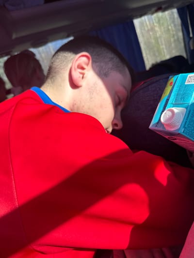 The image shows a person sleeping indoors. The person is lying down, wearing a red shirt with a blue collar. A carton of juice is resting near their head. The text on the carton indicates that it is "Banana-Strawberry" juice. The OCR text implies it might be produced in Uzbekistan (UZ) and intended for the preschool market. The image contains text with dates 28.02.26 and 28.02.21 12:3. The image's location, Челва, Russia, is not visible.
