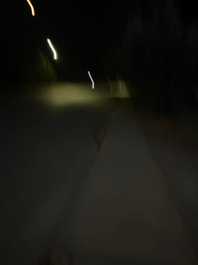 A dark, blurry photograph shows a mix of shadowy and light areas. A tall, brighter section is visible in the middle, suggesting a light source. In the left side of the photo, there are blurred streaks of light, possibly from moving lights. A large, dark wall-like form fills the right portion of the image. The overall impression is one of motion and darkness.