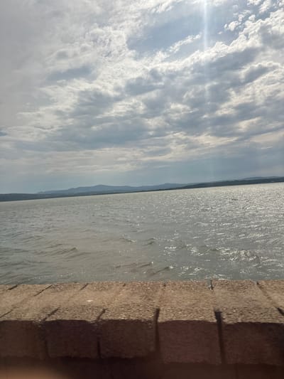 A wide shot shows a body of water, likely a lake or sea, under a cloudy sky with sunlight piercing through. In the foreground, a textured, brown brick wall runs horizontally across the bottom of the frame. The water has small waves and reflects the light from the sky. In the distance, a faint outline of hills or mountains can be seen against the horizon. The scene appears to be outdoors in Port-la-Nouvelle, France, possibly near a waterfront promenade or barrier.