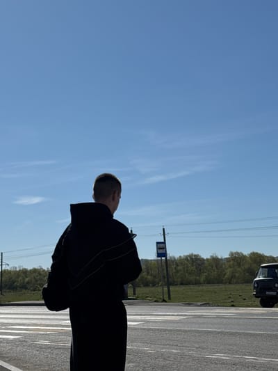 A partially obscured person is standing on the side of a road in Krilovo, Russia. A car is parked on the road. A bus stop is visible with trees in the background, under a bright blue sky with a few faint clouds.