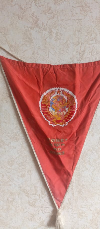 A red triangular pennant hangs on a textured, light-colored wall. The pennant features a circular emblem with a globe, wheat stalks, and a star, which appears to be the coat of arms of the Soviet Union. Below the emblem, gold lettering is visible, though it is partially obscured and difficult to read. A white cord runs along the top edge of the pennant, and a white tassel hangs from the bottom point. The image does not provide any information about the city of Sokol, Russia.