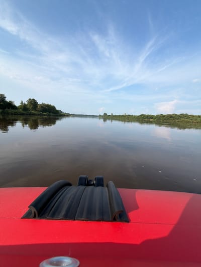The image shows a view from a red boat on a wide, calm river, with lush green trees lining the banks. The sky is blue with wispy white clouds. The foreground is dominated by the red surface of the boat and a black, ribbed structure. The river reflects the sky and the trees. This scene is consistent with the appearance of the Vyatka River in or near the village of Kalino, Russia.