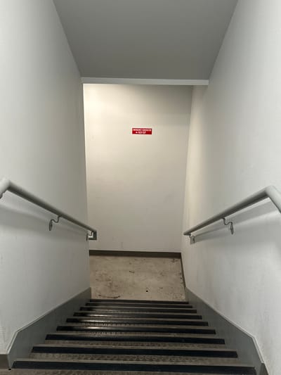 The image shows a view looking down a concrete stairwell with gray metal handrails on both sides. The walls are painted white, and the ceiling is a lighter shade of gray. At the top of the stairwell, a small red sign with white lettering is visible on the far wall, reading "EMERGENCY GENERATOR" with an arrow pointing left and "FACP/SCP". The floor at the bottom of the visible stairs appears to be a concrete landing. The lighting is artificial, casting shadows on the stairs. There are no people or animals in the image. The environment suggests an indoor, likely institutional or commercial building. This image was taken in Lynnwood, United States.