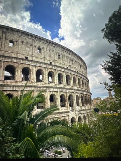 The image depicts a large, multi-tiered ancient Roman amphitheater, commonly recognized as the Colosseum, with the scene located in the city of Kumanovo, North Macedonia. The monumental structure, constructed from weathered light-colored stone, occupies the majority of the left and central frame. It features multiple levels of arched openings, characteristic of Roman architecture, with the uppermost visible tier containing smaller, rectangular window openings and decorative elements.

In the immediate foreground, dense green foliage, predominantly large palm fronds, obscures the lower portion of the amphitheater, particularly on the left side. Additional green trees and shrubs are visible along the right edge of the frame. Through gaps in the foliage at the bottom, a street or pathway is discernible, where indistinct human figures, including one wearing a light-colored shirt, are present. Small white planters with green bushes are also visible at ground level.

In the distant background to the right of the amphitheater, remnants of other ancient stone structures and a tall, slender brown tower rise above the tree line. The sky above is partly cloudy, featuring patches of vibrant blue sky interspersed with white and grey clouds. No specific actions or interactions are clearly detailed among the visible figures.