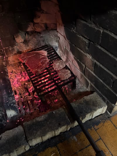 The image shows a grill inside a brick structure, likely a fireplace or oven. The grill contains a large piece of raw meat, possibly a steak, and two smaller circular pieces of meat, possibly burgers or patties. Below the grill are burning coals or embers, creating a red glow and smoke. The brickwork of the structure is visible on the sides and above the grill. A metal rod or handle is visible extending into the scene from the upper right. Location of the scene is unspecified.