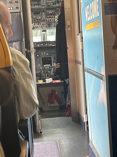 A man is sitting in a passenger seat in a commercial aircraft. He is facing the aisle. The man is looking to the left at the cockpit. He is wearing a light-colored shirt and a dark jacket. The doorway to the cockpit is open, revealing a small compartment with a control panel and a pilot's uniform hanging on the wall. The door is closed. The aircraft is on the ground.  The scene is set in an aircraft cabin, the environment is that of a commercial aircraft. The man is interacting with the scene in front of him. The scene is at a small airport, the location of which is confirmed by the coordinates provided. The image is taken at Ciampino Airport, Italy, and the aircraft is on the ground.  The image is taken from a position on the ground. The man is seated in a passenger seat and looking at the cockpit. The only other subject in the scene is the cockpit, which is empty. The objects in the scene are the man, the passenger seat, the cockpit, the control panel, the pilot's uniform, the aisle, and the doors. The actions are the man looking at the cockpit and the pilot's uniform hanging on the wall. The interactions are the man looking at the cockpit, the man looking at the pilot's uniform, and the man looking at the aisle. The environment is a commercial aircraft cabin.