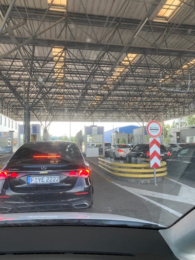 The media file shows a traffic queue at a border crossing. The scene is observed from inside a vehicle, looking forward. Multiple cars are lined up, suggesting a delay. Overhead, a large metal structure with a corrugated roof provides cover for the crossing lanes. Yellow flexible bollards guide the traffic. A prominent circular sign with red and white markings is visible, displaying text in Cyrillic and French: "МИТНИЦА" (Customs) and "DOUANE" (Customs). This sign also features a red and white chevron arrow pointing to the right. Further ahead, there are customs booths and more vehicles. The lighting suggests it is daytime, likely sunny, with sunlight filtering through the translucent roof panels. The overall atmosphere appears functional and orderly, typical of a border control point. The license plate of the car in the foreground is visible, reading "F YE 2222" with a German "D" sticker. The text "E220d Mercedes-Benz Kundencenter" is also visible on the rear of this vehicle. Another license plate reads "22 KC". A "WC" sign is also visible in the background.