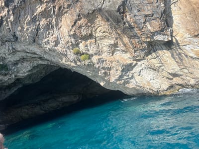 A wide-angle, eye-level shot captures a dramatic sea cave carved into a rugged cliff face, illuminated by sunlight. The cave entrance is a dark, cavernous opening leading into the cliff. The water in front of the cave is a vibrant turquoise blue, with gentle ripples on its surface. The cliff itself is composed of textured rock, showing variations in color from light tan to darker brown and gray. Some patches of green vegetation cling to the upper parts of the cliff. The scene appears to be a natural coastal landscape, likely within the Municipal Unit of Ellomenos, Greece, known for its beautiful coastlines. The overall impression is one of natural beauty and geological grandeur.