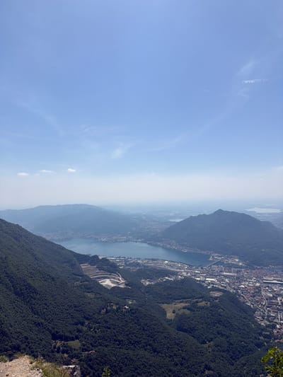 Here's a description of the image:
The image depicts an aerial view of the city of Lecco, Italy, and the surrounding mountainous landscape. The photograph appears to have been taken from a high vantage point, possibly a mountain peak, looking towards the city and Lake Como.
The sky is a pale blue, and there is a haze obscuring the mid-ground.
Lake Como is seen winding between the mountains and the city. The city of Lecco is visible along the shoreline, with its buildings, streets, and industrial areas.
The slopes of the mountains are densely covered with vegetation. A quarry or open mine is visible on one of the slopes.