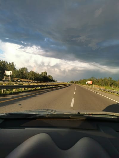 The image shows a view from inside a vehicle traveling on a highway. The highway has multiple lanes and is bordered by a guardrail and trees. The sky is dramatic, with dark storm clouds in the upper portion and brighter, lighter clouds towards the horizon, where a faint rainbow is visible. A sign with the word "MORT" is visible on the right side of the road. The dashboard of the vehicle is in the foreground, partially obscuring the view of the road ahead. The scene appears to be in Mercier, Canada, based on the provided information, likely during or just after a rain shower.