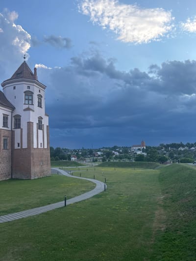 A stone and brick tower, part of a larger building, stands in the foreground to the left of the image. A paved pathway curves through a grassy area with scattered small yellow flowers and low lampposts. In the middle ground, a town with houses and a prominent church with a red roof is visible. The background is dominated by a dramatic sky with dark, heavy clouds contrasting with patches of brighter blue sky and white clouds. Two birds are visible in the sky. The overall scene suggests a historic location on a somewhat overcast day with a sense of impending weather change.