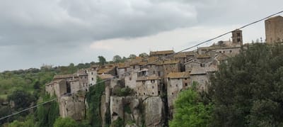 The image depicts Vitorchiano, Italy, a town built on rocky cliffs. Buildings, constructed of stone with terracotta tiled roofs, line the cliff edge. A church bell tower and a medieval tower are visible among the rooftops. Green foliage, including trees and ivy, grows on the cliffs below the buildings. Overhead, grey, overcast sky with low clouds. A power line diagonally crosses the frame.