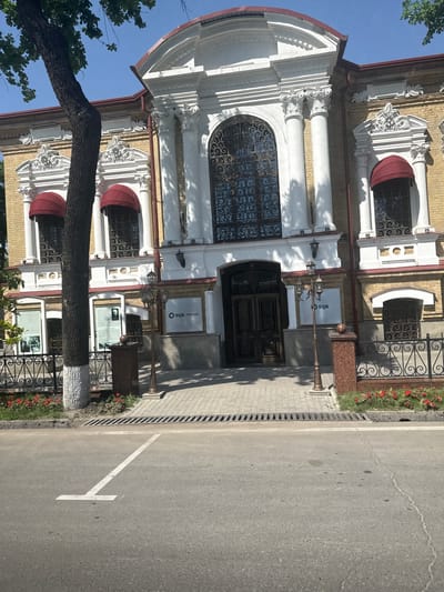 The image shows the facade of a building, likely a commercial or public institution, in Kashgar mahalla, Uzbekistan. The architecture is ornate, with white columns, intricate detailing around windows, and a rounded top section. Red awnings are above some windows. The building is constructed of light-colored brick. A dark-colored, possibly metal, door is visible at the entrance, flanked by signage for 'SQB Premium'. A decorative lamppost is to the side of the door. A tree trunk is in the foreground, partially obscuring the building. The street adjacent to the building is paved, with a white parking space marking visible. A strip of red flowers grows along the building's foundation behind a low, decorative iron fence.