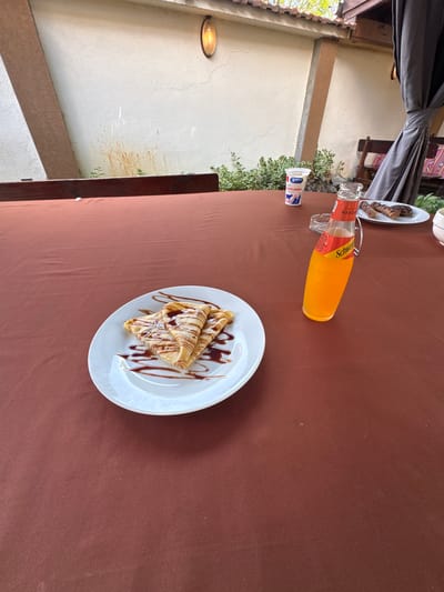 The image displays a close-up, slightly overhead view of a table in an outdoor or semi-outdoor dining setting. A dark brown tablecloth covers the table surface.

In the foreground, a white plate presents two triangular-folded crepes or pancakes, generously drizzled with chocolate syrup and lightly dusted with powdered sugar or white shavings. Beside the plate, a clear glass bottle containing an orange liquid lies on its side. Its label prominently features "Schweppes" and "MANGO," identifying it as a mango-flavored drink. The bottle has a distinctive glass handle. Near the bottle, a small white plastic cup with blue and red branding is visible, with "MILKO" printed on its label. In the upper left, a portion of another white plate is visible, holding several pieces of what appear to be grilled meat or sausages.

The background elements suggest an open-air or veranda-style environment. Dark grey curtains or drapes are tied back, revealing a light-colored wall with visible cracks and some brown staining. A wooden beam runs horizontally across the wall. Green foliage from plants or bushes peeks out from behind the table edge near the wall. An oval-shaped outdoor light fixture, emitting a warm glow, is mounted on the wall in the upper left.

The scene depicts a casual daytime meal or snack. No individuals are present in the image. The setting is likely a restaurant or cafe patio in Gorna Grashtitsa, Bulgaria.