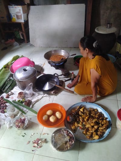 A woman in a yellow garment is sitting on the floor in a room, likely a kitchen, preparing food. The environment appears to be a modest living space, possibly in Kruengbaroblangme, Indonesia, given the context. She is situated to the right of the frame, facing away from the viewer. In front of her, on the tiled floor, are various ingredients and cooking utensils. A large pot with a reddish-brown mixture is on a stovetop in the center of the image, suggesting active cooking. Other items include a pink basin, a metal pot with a lid, a wok, a bowl of potatoes in broth, a platter of cooked chicken, and raw meat in a small bowl. There are also fresh ingredients like shallots, garlic, limes, and banana leaves scattered around. The lighting suggests it is daytime, with natural light entering the room. The overall scene depicts a moment of food preparation in a domestic setting.