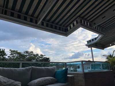 The image depicts an elevated outdoor patio or balcony in Floridablanca, Colombia, offering a view of a residential area and distant natural landscape under a cloudy sky. In the left foreground, a section of a light gray outdoor sofa is visible, furnished with multiple cushions, including one teal-colored accent pillow. A glass railing with a base constructed of horizontal light-colored planks extends across the mid-ground, separating the patio from the exterior view. Overhead, a distinctive ceiling or awning structure is present, characterized by alternating light and dark parallel stripes. This structure features an angled cut-out, exposing a patch of cloudy sky, and includes a diagonal support beam on the right, also adorned with the striped pattern.
Beyond the railing, a dense canopy of green trees forms a significant portion of the mid-ground. Below the treeline, several residential buildings with red-tiled roofs and light-colored walls are discernible. In the background, beyond the residential structures, the faint outlines of hills or mountains are visible under a sky predominantly filled with large, white and gray cumulus clouds. A palm frond is partially visible on the far right. No individuals, specific actions, or interactions are present within the frame. The scene is illuminated by diffused daylight, consistent with overcast or partially cloudy conditions.