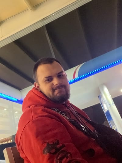 The scene depicts a single male subject positioned in the foreground at an outdoor commercial establishment, consistent with a gas station, located in Piperkov Chiflik, Bulgaria. The individual, appearing to be an adult male, has short, dark hair and a full beard and mustache. He is wearing a red hooded sweatshirt, which features dark graphic designs on the sleeves. A white-colored lanyard with the text "MCYSF" is visible around his neck, and a black strap, possibly from a bag or camera, crosses his chest. His gaze is directed towards the viewer, with a neutral facial expression.

The background consists of a brightly illuminated white canopy structure, under which a blue LED strip light runs horizontally. Above the canopy, a darker, possibly recessed ceiling is visible. To the subject's right, a white support pillar is present, displaying the number "21" in a dark color. The overall lighting suggests artificial illumination, characteristic of a nighttime or covered environment. No other individuals or specific actions are discernible.