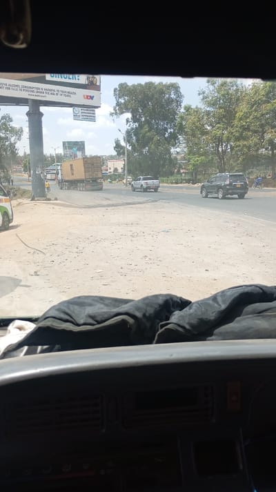 A street scene is viewed from inside a vehicle's dashboard. In the background, a large billboard is visible with an advertisement that includes the text "EXCESSIVE ALCOHOL CONSUMPTION IS HARMFUL TO YOUR HEALTH. NOT FOR SALE TO PERSONS UNDER THE AGE OF 18 YEARS" along with a website address. The advertisement also displays phone numbers. In the distance, traffic is visible, including a truck, SUV, and pick-up truck. Trees line the horizon, and the ground is dirt and gravel.
